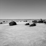 black and white photo of beach with wavy sand and rocks silhouetted